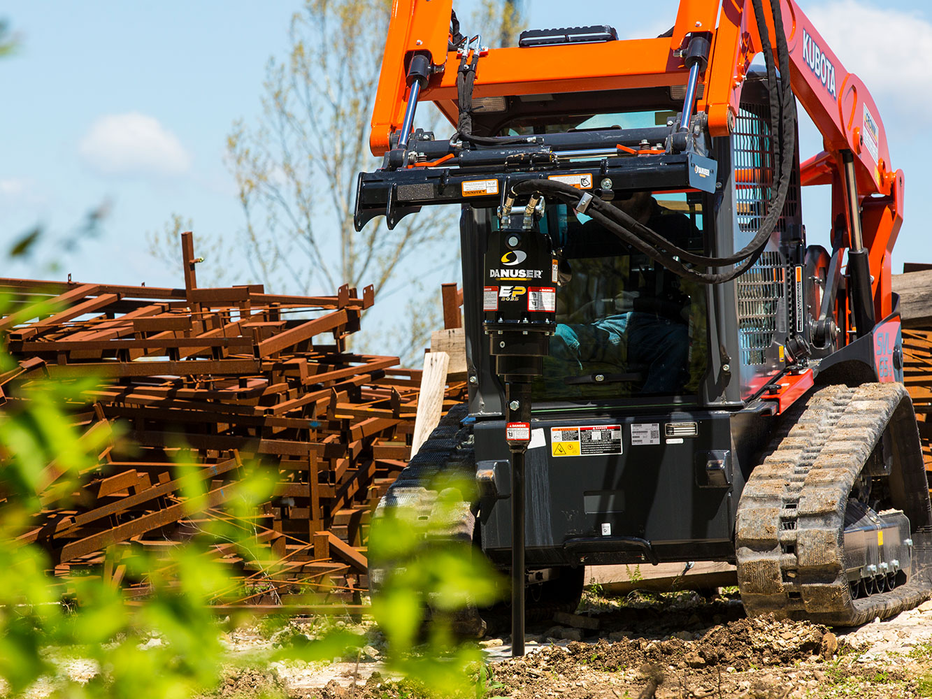 Image showing Danuser EP 2035 Auger Drive on Kubota SVL 65-2 Skid Steer near scrap metal pile, trees, and cloudy sky on uneven ground