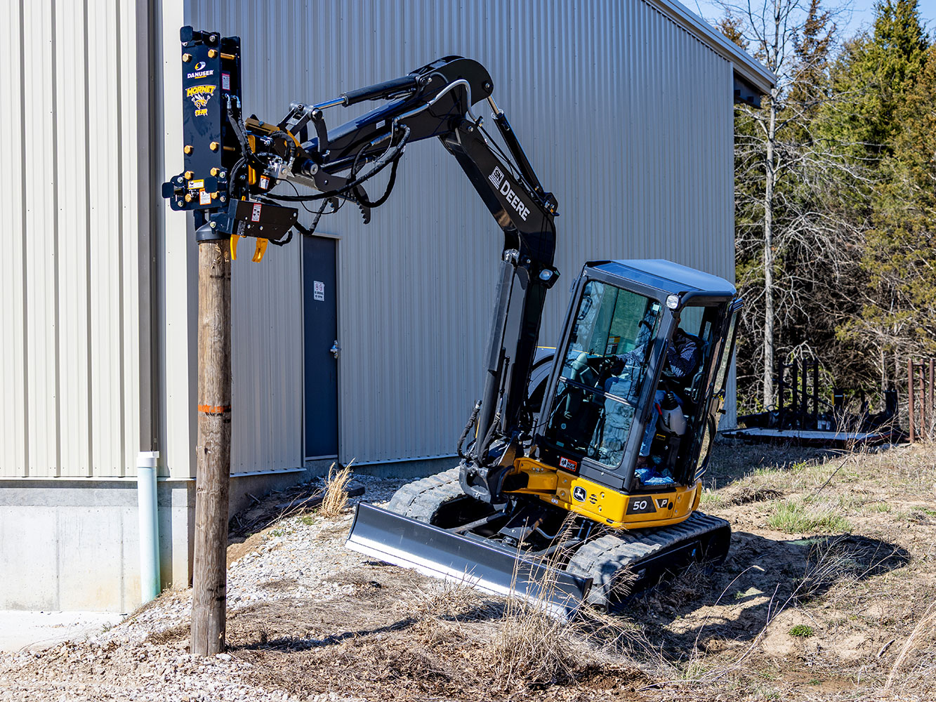 Image showing Danuser Hornet T8-RR post driver on Mini John Deere 50P Excavator holding wood post beside building using tilt feature