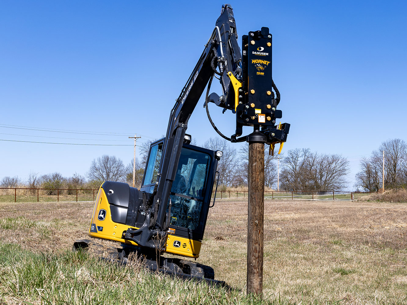 Image shows Danuser Hornet T8-RR post driver on a Mini John Deere 50P Excavator drives a wood post into the ground on a hillside