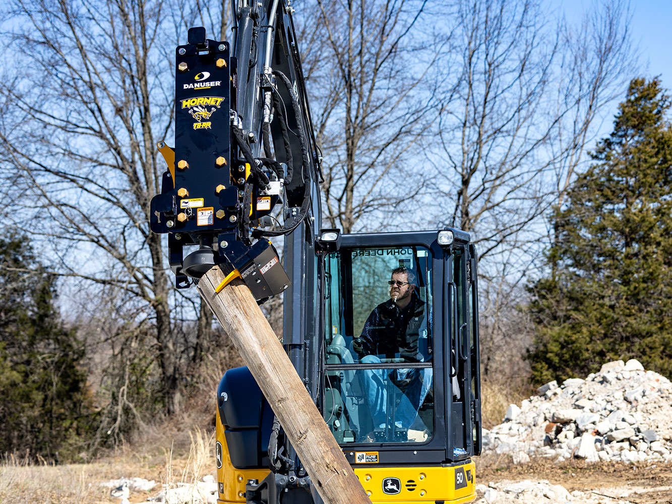 Image showing Danuser Hornet T8-RR post driver on Mini John Deere 50P uses grapple to move wood post in field, preparing for installation