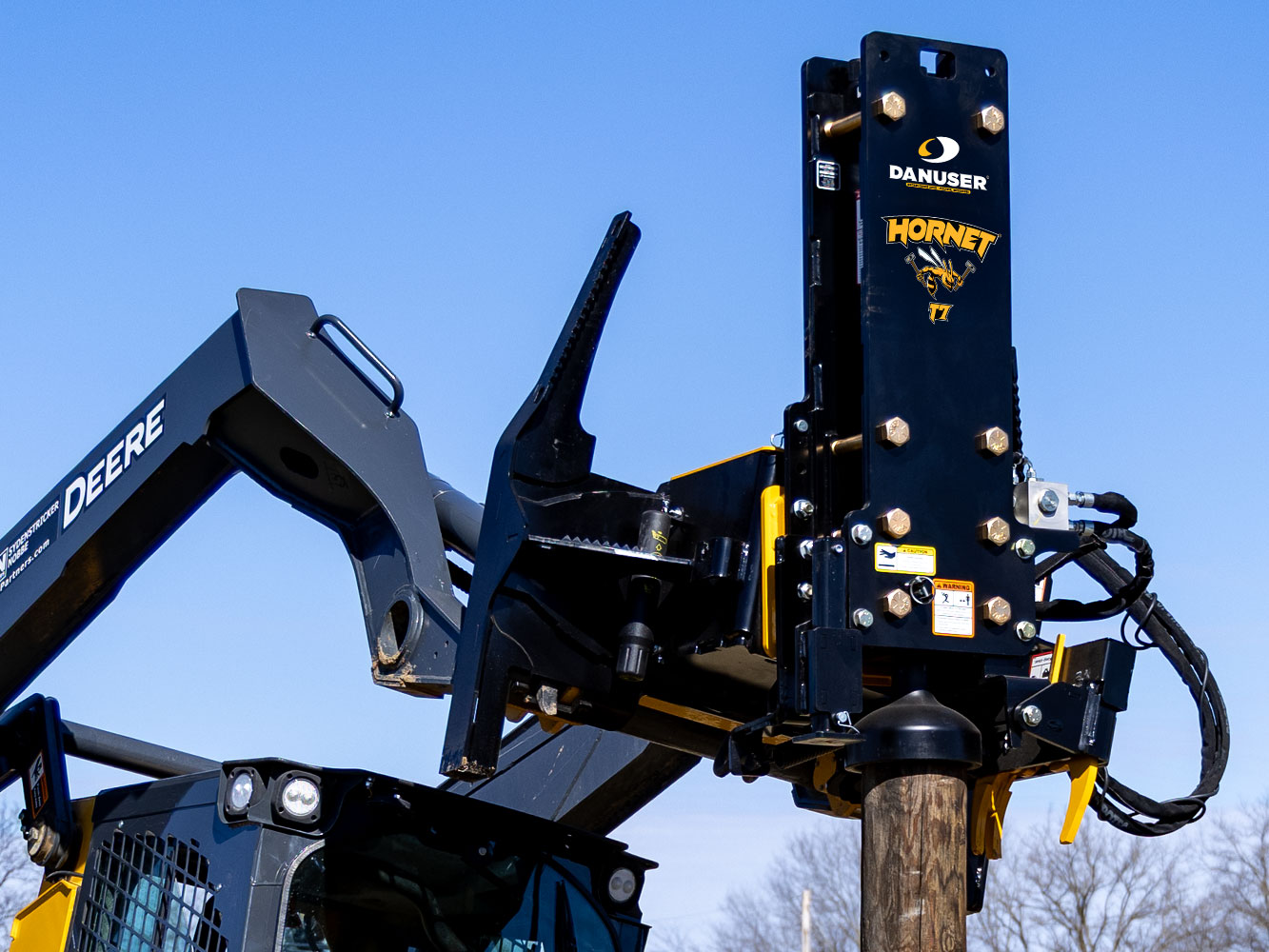 Image showing close-up of Danuser Hornet T7 post driver on John Deere 333G Skid Steer with wood post, blue sky, and tree tops