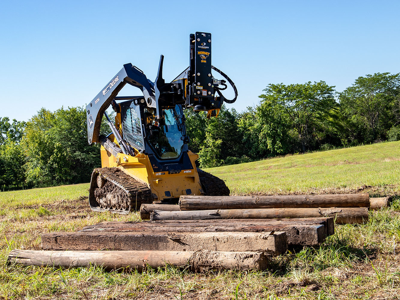 Image shows Danuser Hornet T8-RR on John Deere 333G Skid Steer in grassy field behind pile of wooden posts with trees in background