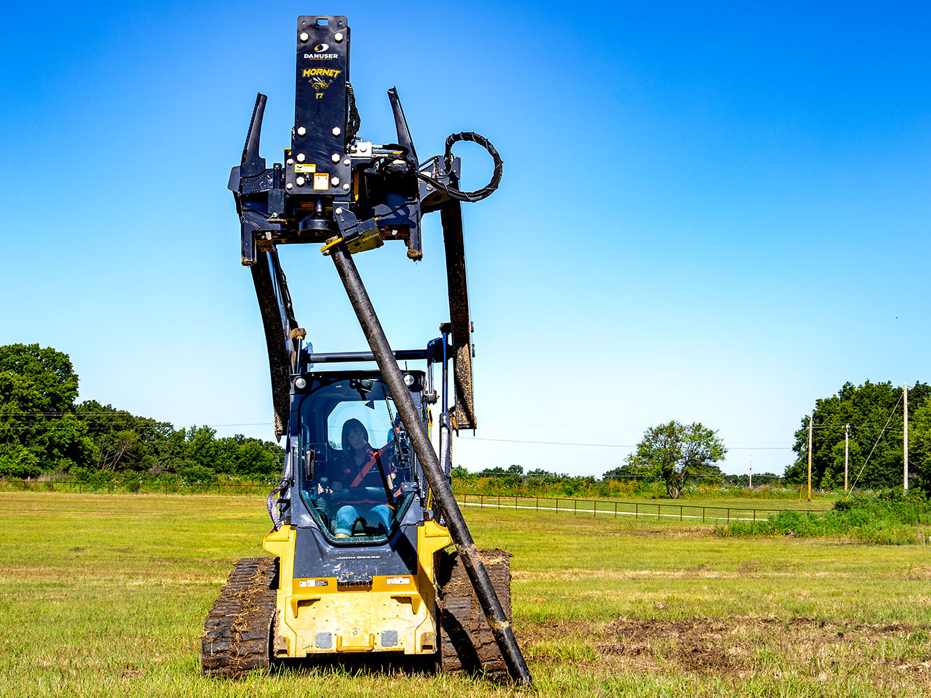 Image shows Danuser Hornet T7 on John Deere 333G Skid Steer using grapple to hold tilted post in grassy field during post installation