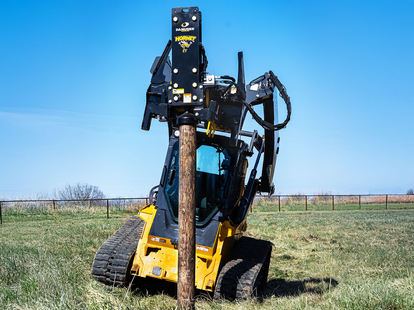 Image showing Danuser Hornet T7 with tilt feature on John Deere 333G Skid Steer driving wooden post into ground in open field with blue sky