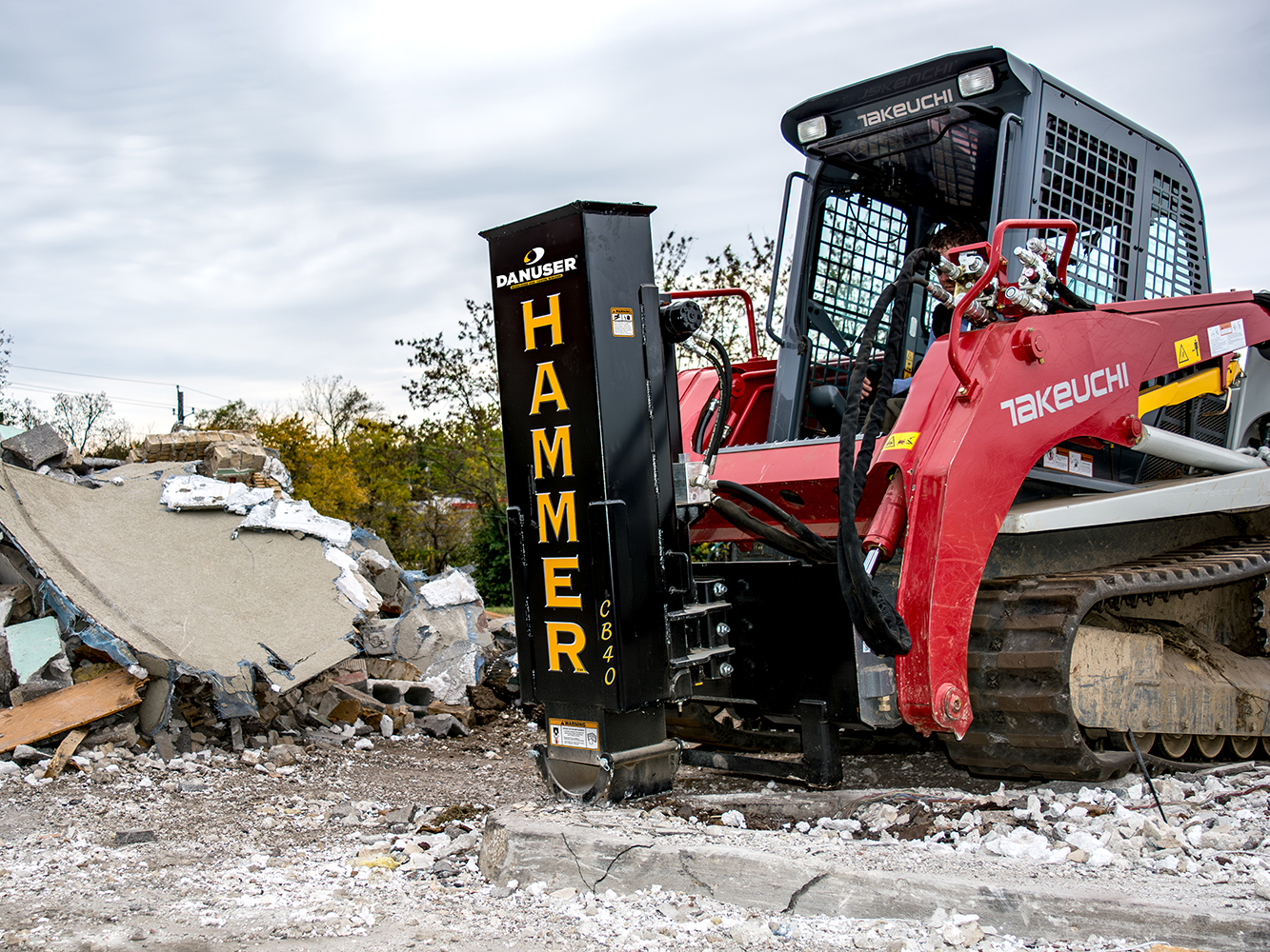 A red Takeuchi skid steer equipped with a Danuser Hammer CB40 hydraulic breaker attachment at a demolition site, surrounded by rubble and broken concrete.