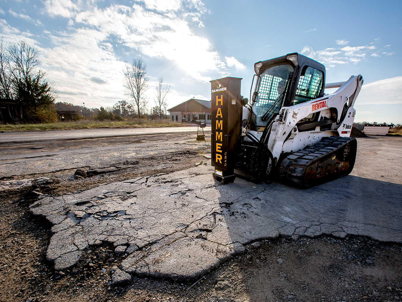 A compact track loader equipped with a Danuser Hammer CB40 hydraulic breaker attachment breaks up cracked asphalt in an open area near trees and a building.