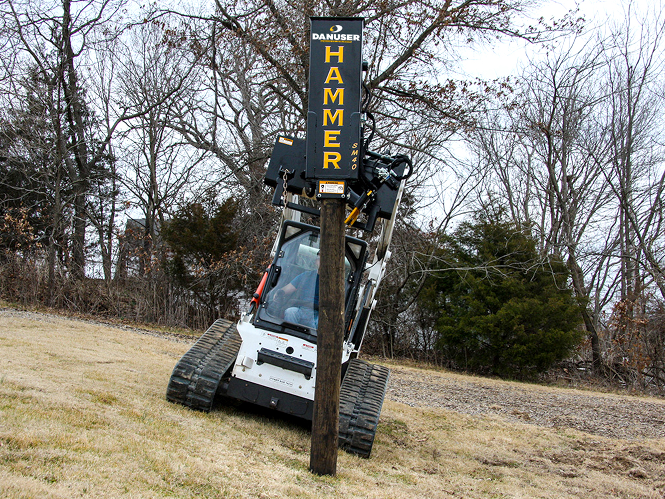 A skid steer loader equipped with a Danuser Hammer SM40 post driver attachment with tilt is positioned on a grassy hill, driving a wooden post, with dense green foliage in the background.