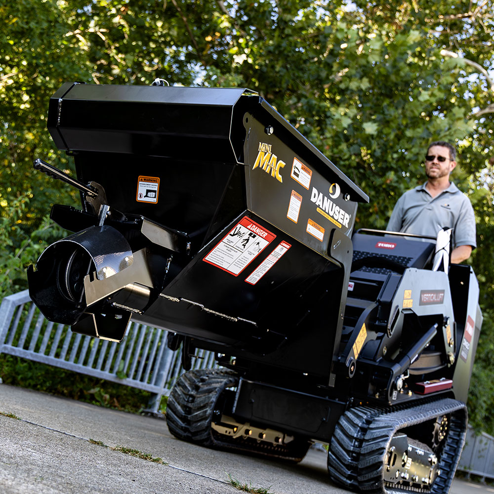 A Danuser Mini Mac material handling bucket mounted on a Toro mini skid steer, positioned on a concrete surface with green foliage in the background. A person operates the skid steer. The bucket displays warning labels and branding, including "Mini MAC" and "Danuser."