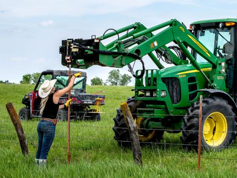 Image showing Woman holds T-post under Danuser T3 on John Deere loader as tractor driver prepares to drive post in fence line