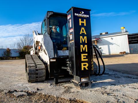 A skid steer loader equipped with a Danuser Hammer CB40 hydraulic breaker attachment breaks up concrete at a construction site, with a building and trees in the background.