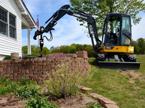 A Deere 35G mini excavator positioned on grass beside a raised brick garden bed near a house, with its arm extended over the bed containing plants and flowers. Trees and an American flag mounted on the house are visible in the background.