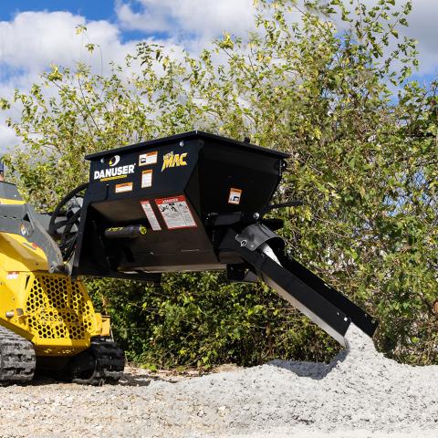 Mini MAC material handling bucket attached to a yellow skid steer loader, dispensing dry concrete onto a pile. The bucket is black with “Danuser” and “Mini MAC” branding visible on its side. Green foliage and a partly cloudy sky appear in the background.