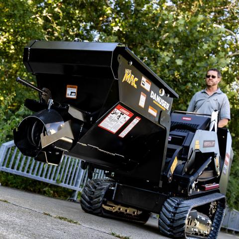 A Danuser Mini Mac material handling bucket mounted on a Toro mini skid steer, positioned on a concrete surface with green foliage in the background. A person operates the skid steer. The bucket displays warning labels and branding, including "Mini MAC" and "Danuser."