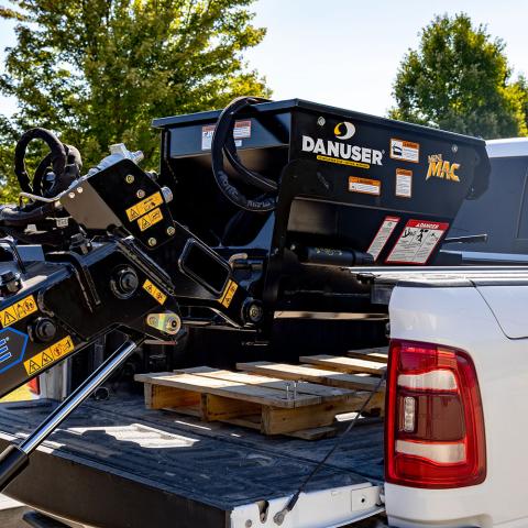 A Danuser Mini Mac material handling bucket being lifted from the bed of a white truck. The black bucket features visible "Mini Mac" and Danuser branding. The truck bed contains wooden pallets, with trees and a clear sky in the background.