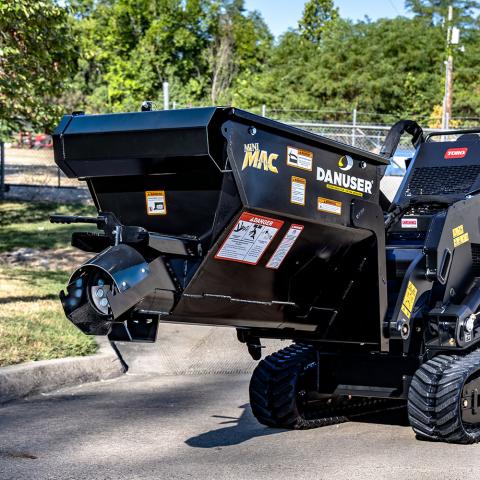 A Danuser Mini Mac material handling bucket mounted on a mini compact track loader. The black bucket features prominent "Mini MAC" and "Danuser" logos, along with multiple warning and instruction labels. The background includes grass, trees, and a chain-link fence.