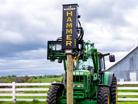 A green tractor with a Danuser EM40 Hammer attachment driving a wooden post into the ground near a white fence in a grassy field.