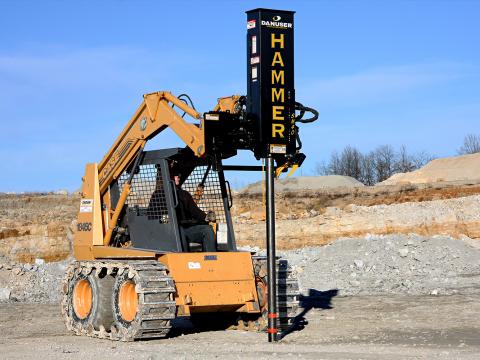 A yellow skid steer loader with a Danuser SM40 Hammer attachment driving a post into rocky ground at a construction site.