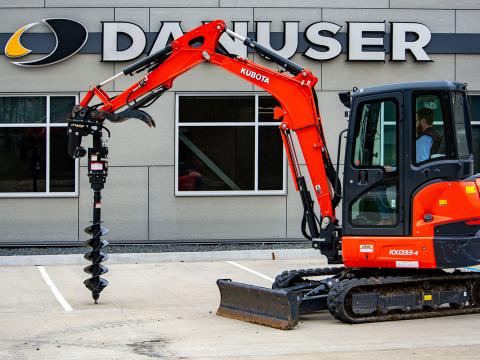A Kubota KX033‑4 mini excavator equipped with a Danuser EP1020 auger drive, positioned on a concrete lot in front of the Danuser office building.